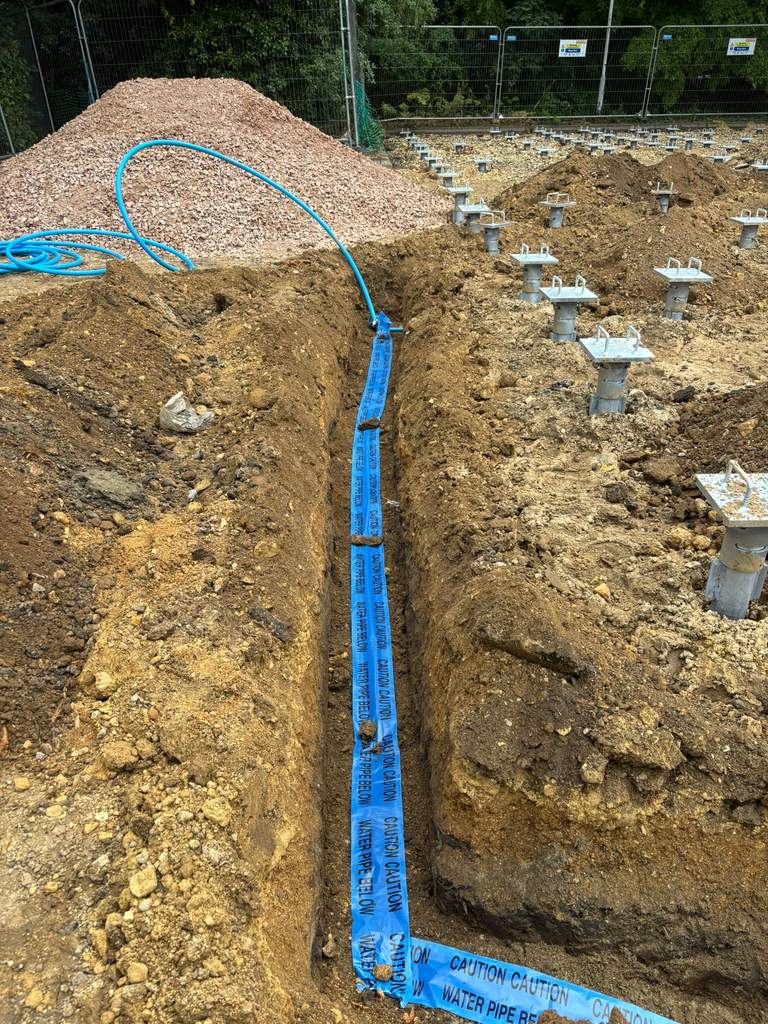 New water main trench with marker tape beside pile foundations